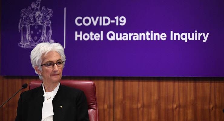 Jennifer Coate sitting at desk in front of hotel quarantine inquiry sign